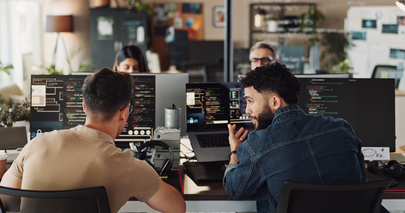 Two developers reviewing code together on a laptop in an open studio, a third at work behind them.
